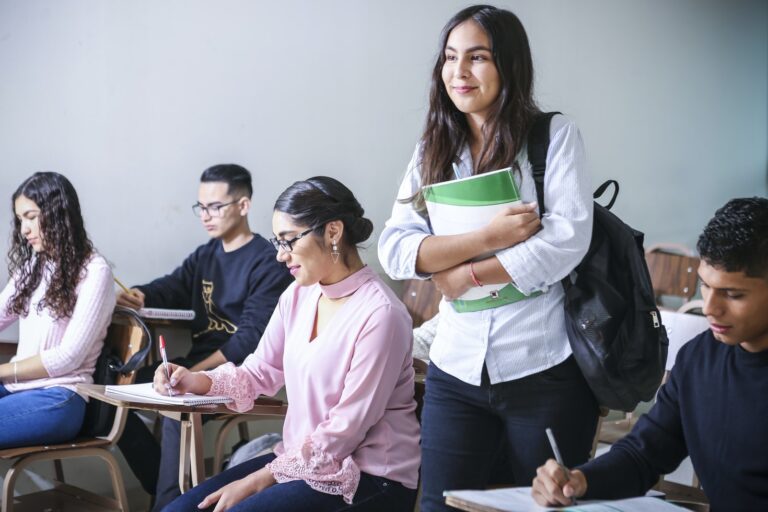 Grupa studentów podczas zajęć w sali wykładowej. Kilka osób siedzi przy ławkach i robi notatki, a młoda kobieta stojąca między nimi trzyma zeszyt i ma na ramieniu plecak.