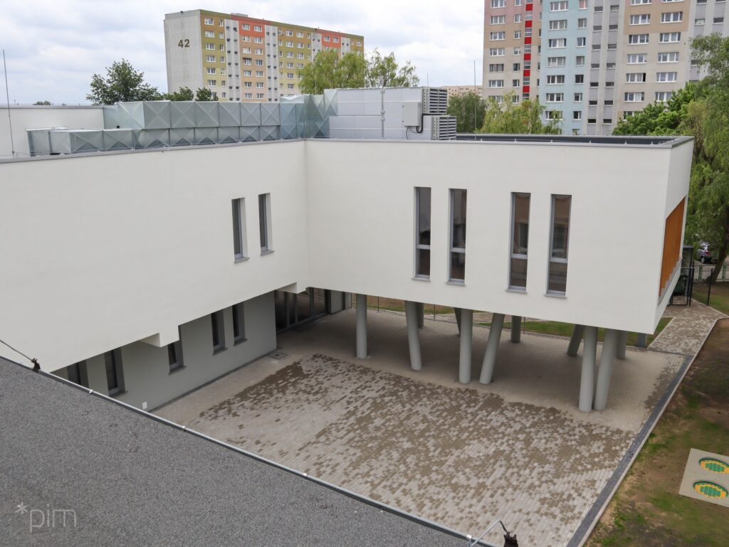 Na zdjęciu budynek sali gimnastycznej, biała elewacja, prawa część budynku posadowiona jest na betonowych palach