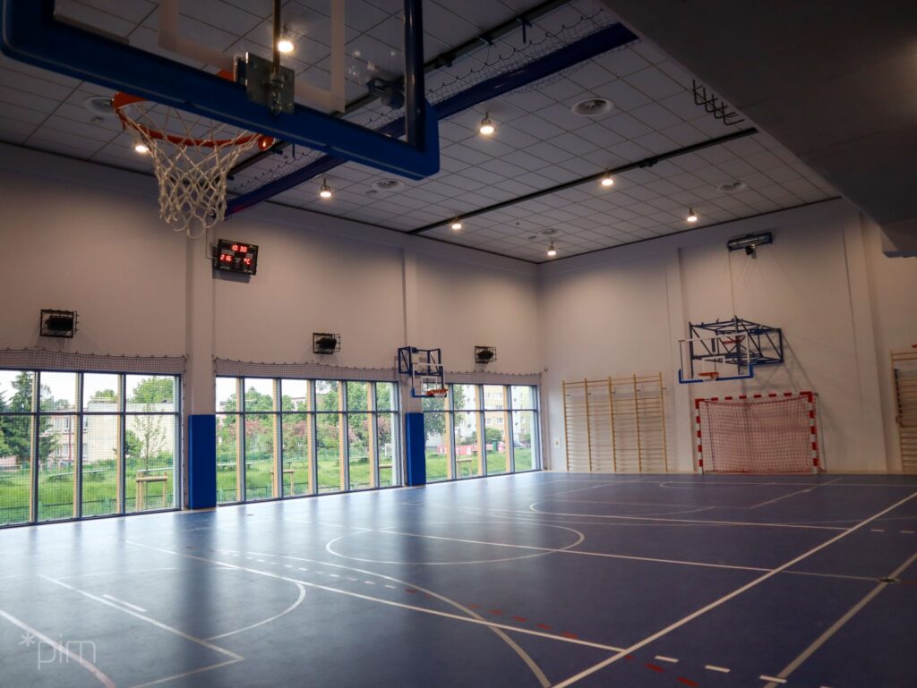 Na zdjęciu wnętrze sali gimnastycznej, na wprost bramka z czerwoną siatką, nad nią kosz do koszykówki, po lewej stronie 3 segmenty dużych przeszklonych okien, pomiedzy dwama segmentami kosz do koszykówki, po przeciwnej stronie trzeci kosz do koszykówki