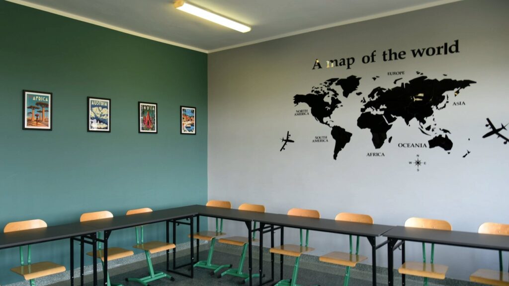 Sala szkoły podstawowej nr 77. Na zdjęciu szare stoły tworzące kat prosty, przy nich drewniane krzesła z metalowymi, jasnozielonymi ramami. Na zielonej ścianie po lewej stronie cztery kolorowe zdjęcia w czarnych ramkach, na szarej ścianie po prawej stronie mapa świata z czarnymi płaszczyznami kontynentów