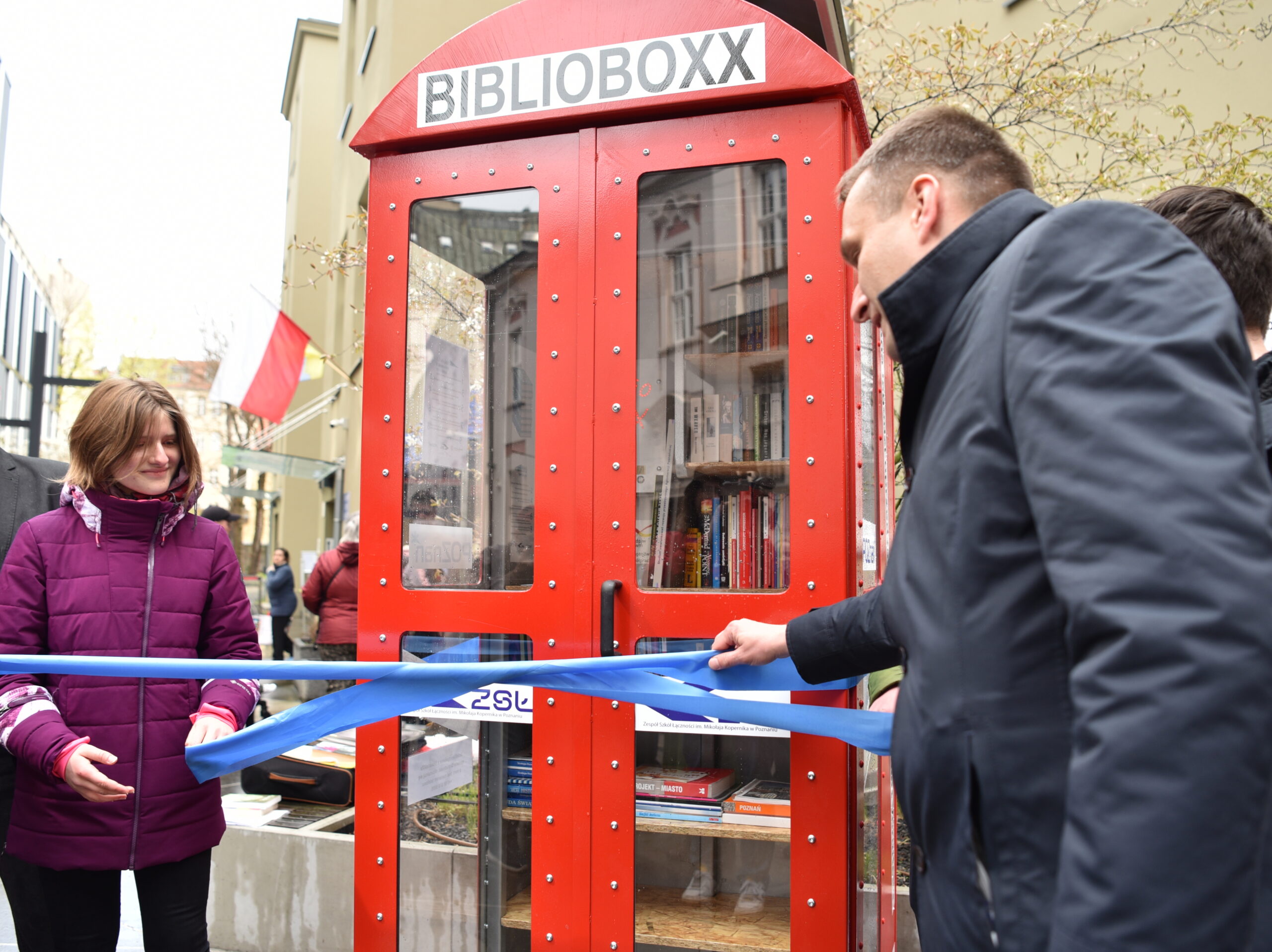 Wiceprezydent Mariusz Wiśniewski rozwiązujący niebieską szarfę w geście otwarcia BiblioboXXu, który wygląda jak londyńska, czerwona budka telefoniczna 