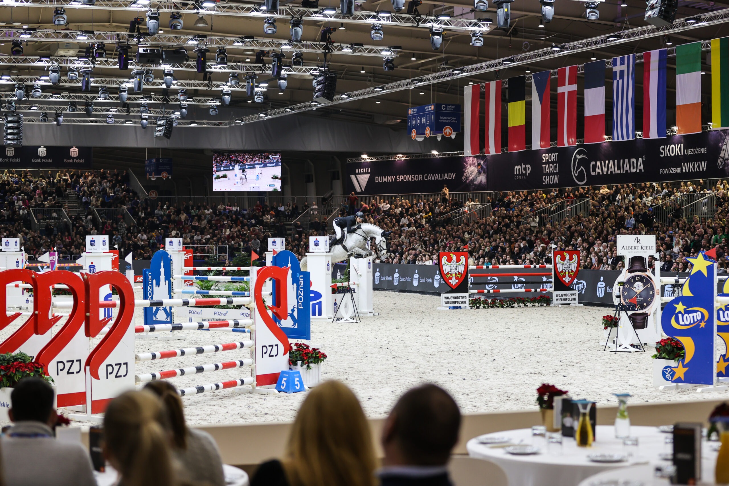 Na zdjęciu zawody konne, jeździec na koniu w trakcie wykonywania skoku przez przeszkodę, na trybunach publiczność, nad trybunami flagi różnych państw