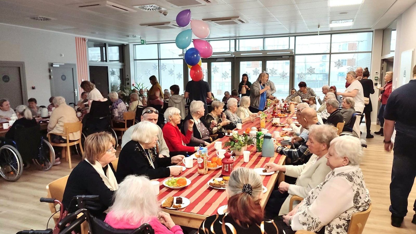 Na zdjęciu stół zastawiony jedzeniem, przy którym siedzą starsze osoby
