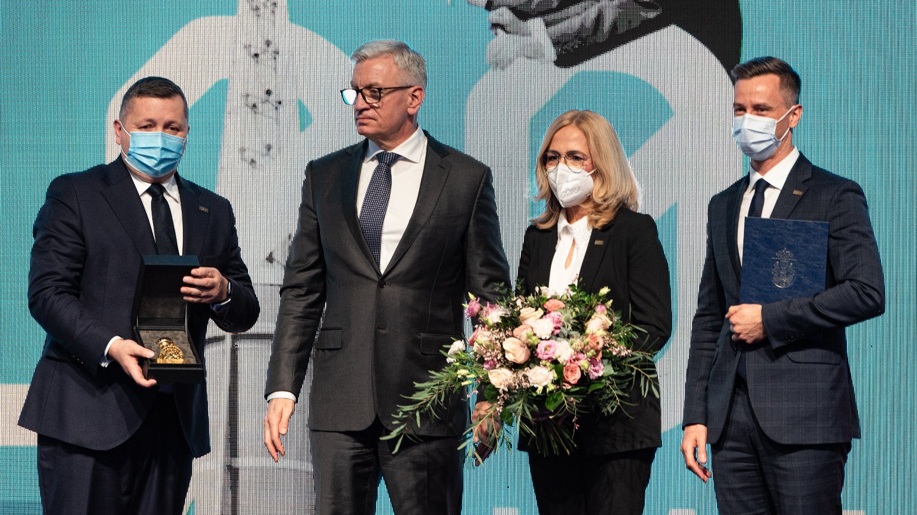 Jacek Jaśkowiak prezydent Poznania stojący obok Tomasza Kobierskiego prezesa Grupy MTP który trzyma w rękach Złotą Pieczęć. Obok prezydenta kobieta w maseczce trzymająca bukiet kwiatów, a obok niej mężczyzna w maseczce z dyplomem