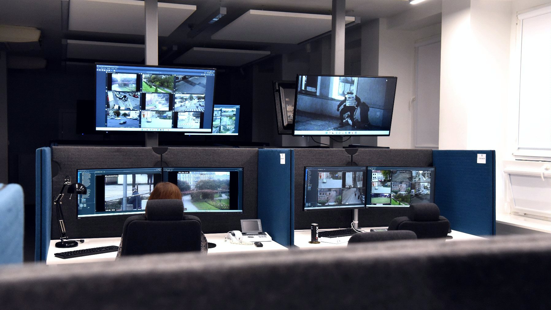 sala wypełniona monitorami przy stanowiskach pracy. Przy jednym z biurek siedzi pracownik patrzący na ekran 