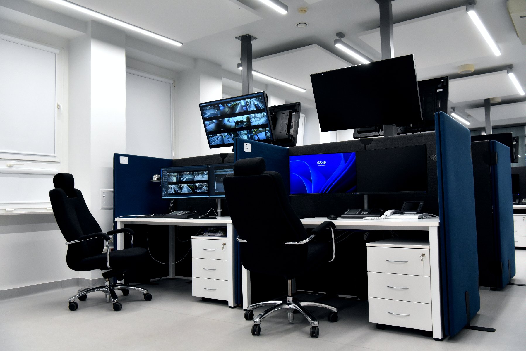 widok na dwa stanowiska monitoringu: dwa białe biurka, dwa czarne fotele oraz 6 monitorów