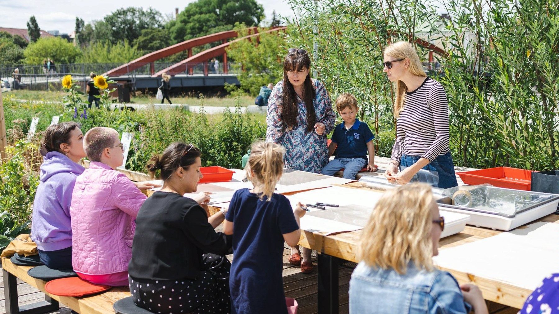 Dzieci i kobiety przy stole w trakcie zajęć plastycznych na świeżym powietrzu, w tle rzeka i most