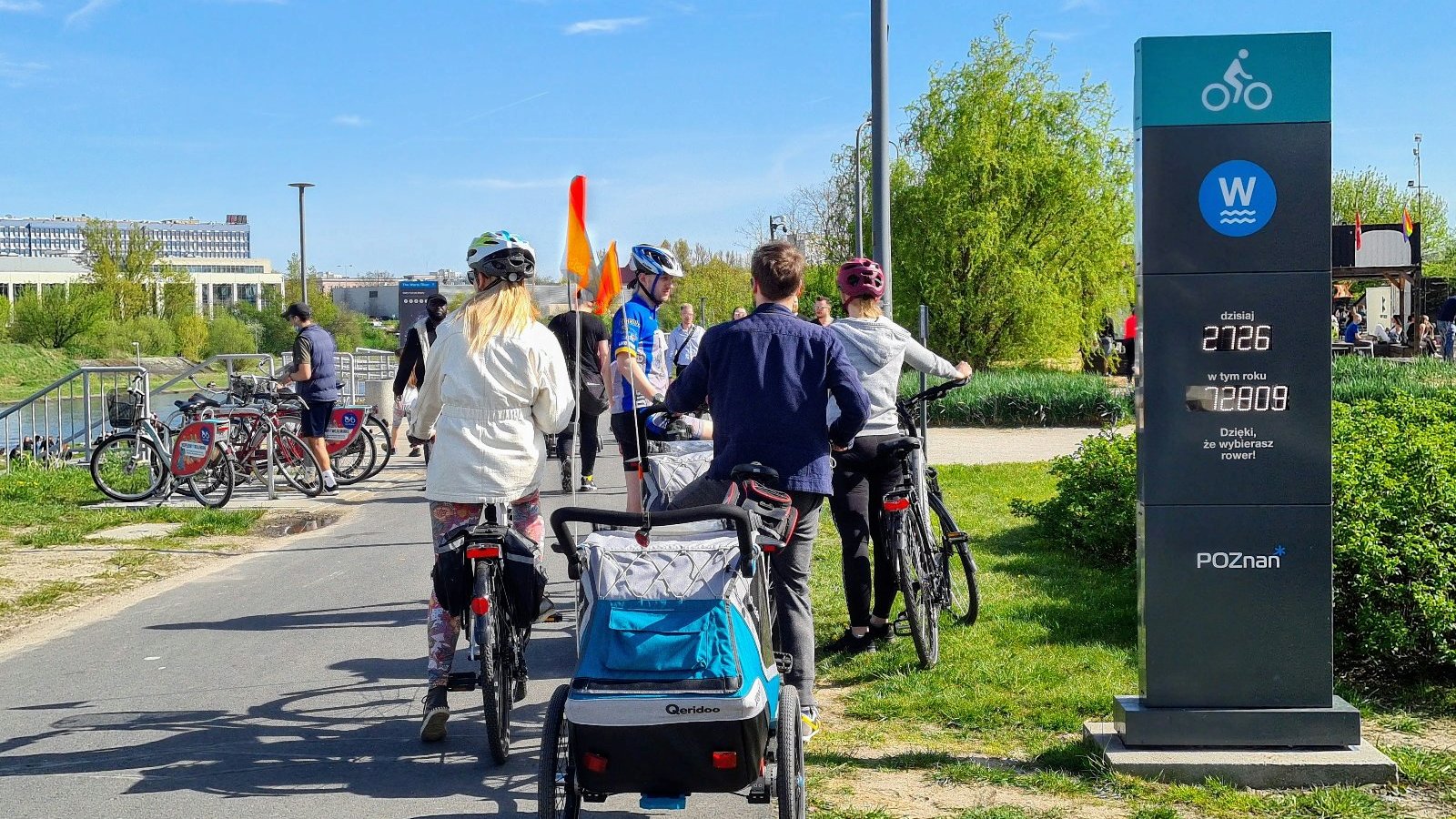 Droga rowerowa nad rzeką Wartą, a na niej ludzie na rowerach i z przyczepką z dziećmi. Ludzie noszą kaski. Na pierwszym planie licznik rowerowy.