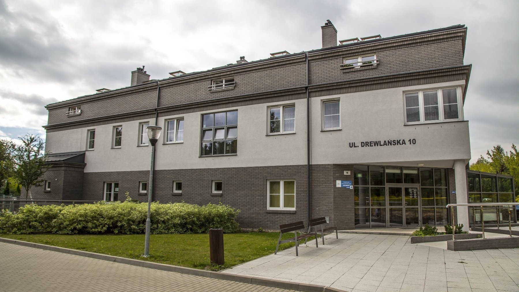 Budynek w kolorze jasnej i ciemnej szarości z adresem "ul. Drewlańska 10" oraz ławki i zieleń przed budynkiem