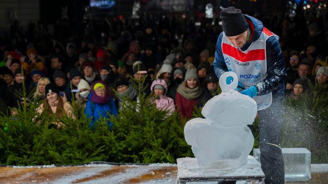 mężczyzna rzeźbiący piłą w lodzie, pod sceną publiczność i rząd choinek