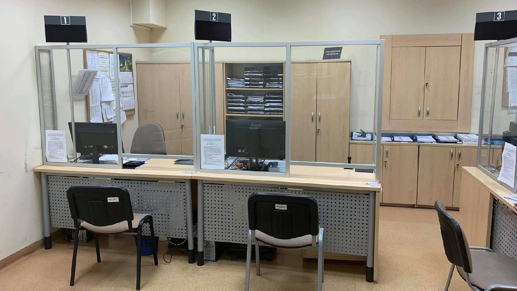 Dwa stanowiska obsługi klienta oddzielające go z pracownikiem pleksą z komputerami. Z tyłu szafki oraz dokumenty.