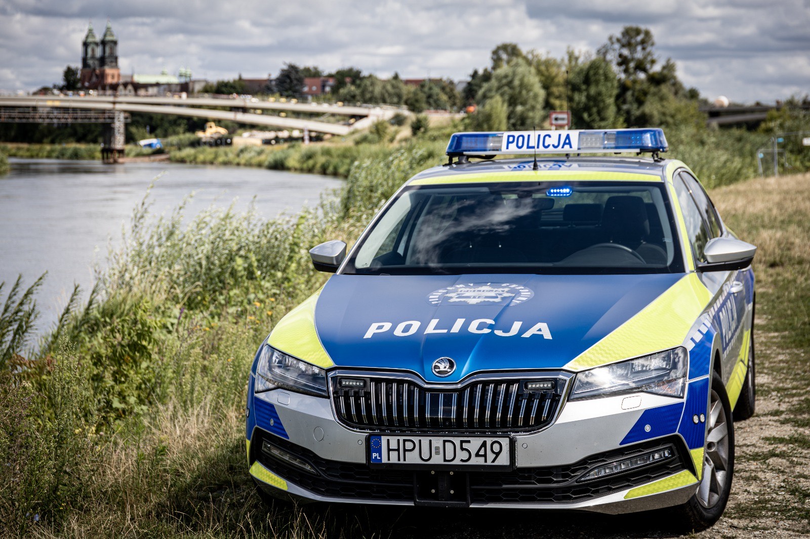 Zielono-srebrno-żółty samochód policji nad brzegiem Warty w tle Ostrów Tumski