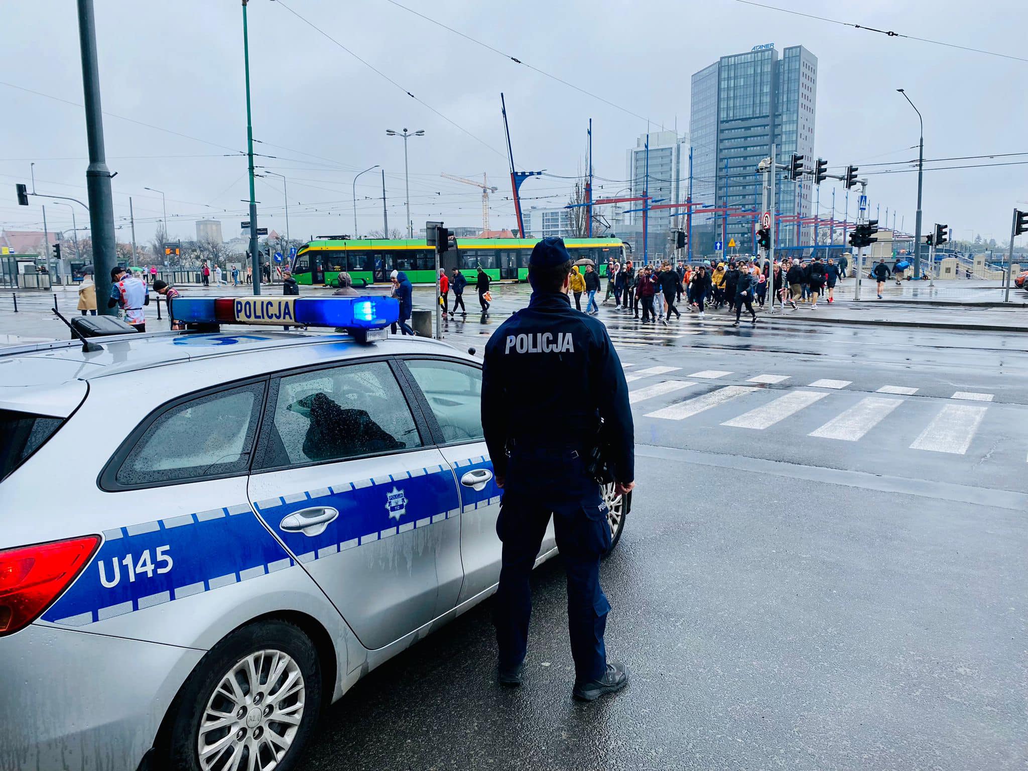 Policjant stoi przy radiowozie i obserwuje ludzi przechodzących na pasach, w tle przejeżdża tramwaj