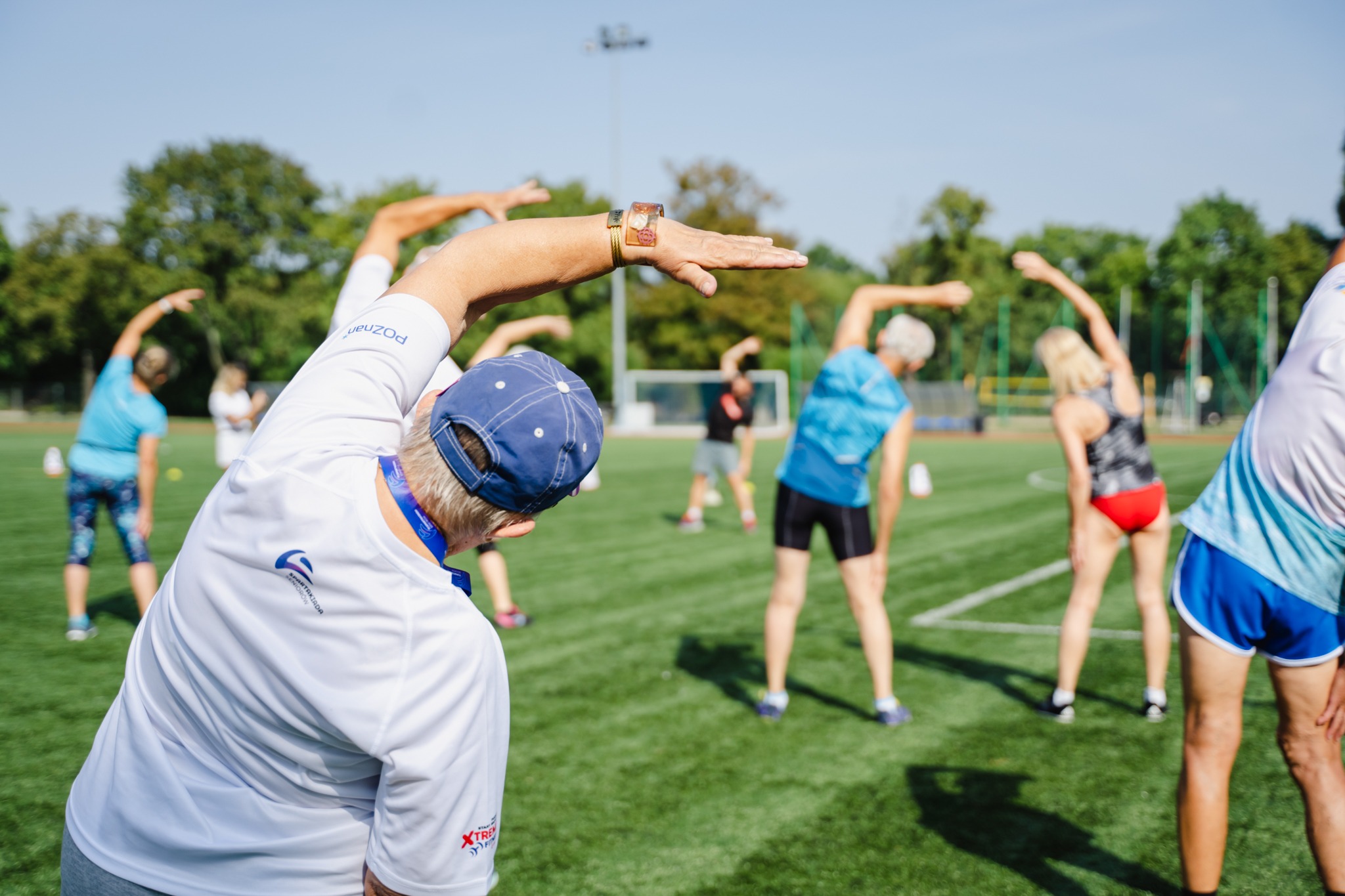 Grupa seniorów uczestniczy w rozgrzewce na boisku. Wykonują skłony boczne w pozycji stojącej.