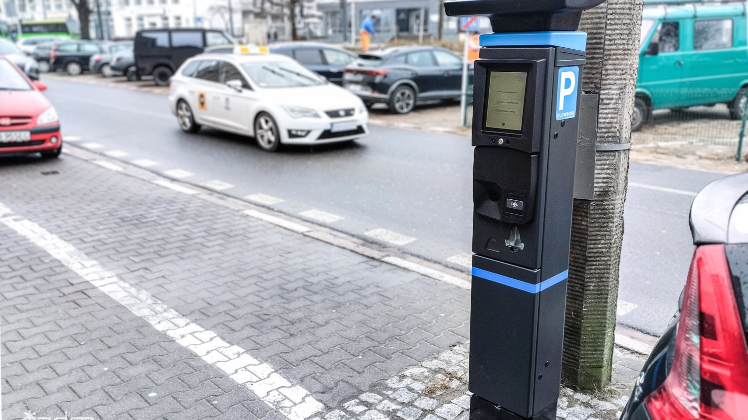 Parkomat stojący na chodniku przy ulicy. Zaparkowane samochody i taksówka jadąca w tle.