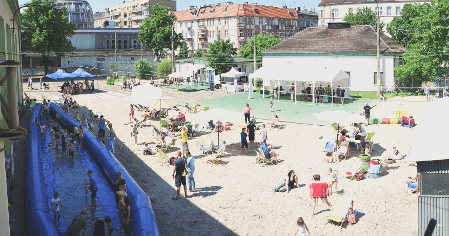 lato na Madalinie - dzieci i dorośli bawiący się i odpoczywający na strefie plażowej z torem wodnym. Namioty i parasole słoneczne. Wokół budynki i intensywne słońce oświetlające okolicę.