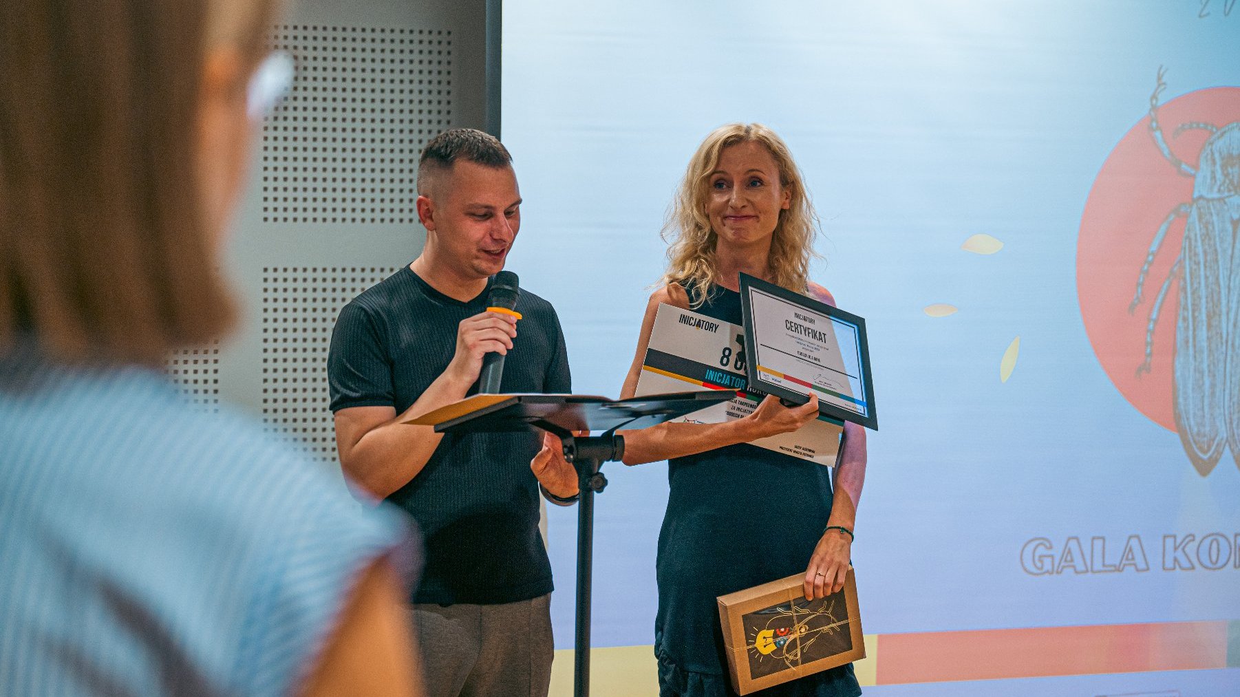 Mężczyzna mówi przez mikforon przy mównicy. Obok kobieta w blond włosach z nagrodą i certyfikatem Inicjatora Roku.