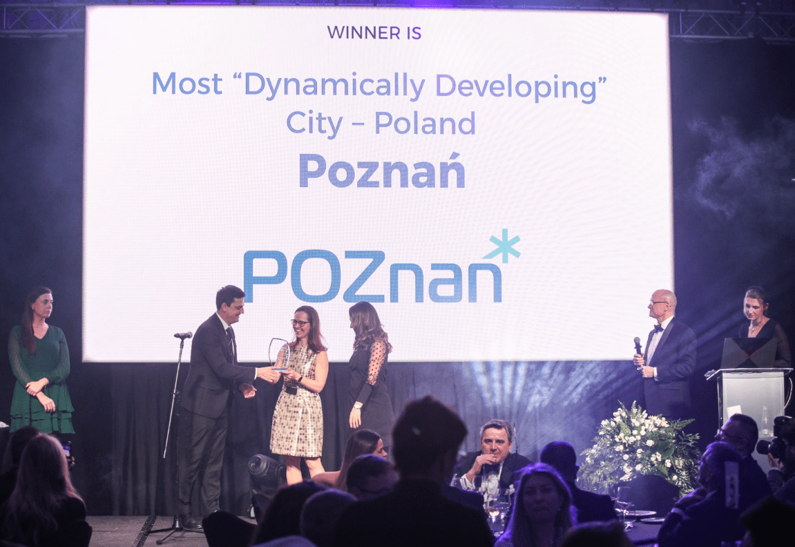 Kobieta odbiera nagrodę na scenie. W tle prezentacja z napisem "Winner is most dynamically developing city - Poland -Poznań, ludzie na widowni i na bokach sceny ubrani w eleganckie ubrania