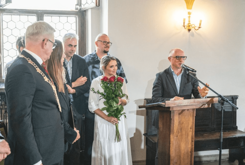 Mężczyzna w garniturze przemawia na mównicy. Obok grupa ludzi. Kobieta w białej sukience trzyma bukiet róż. Obok prezydent Poznania Jacek Jaśkowiak.