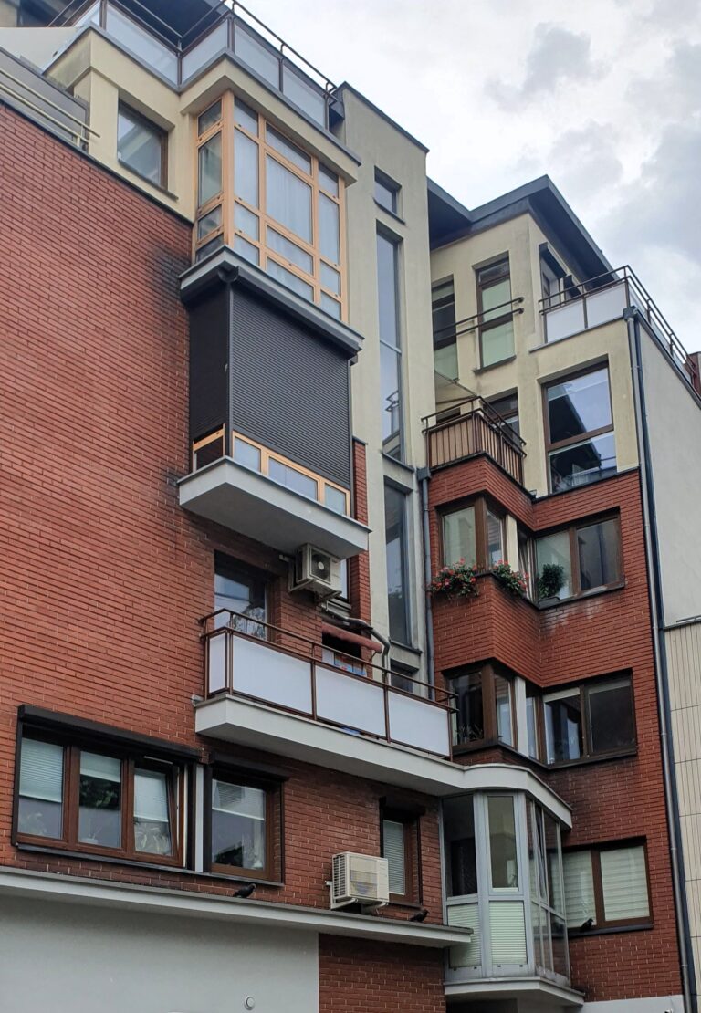 Nowoczesny, wielokondygnacyjny blok mieszkalny z kontrastem czerwonej cegły i jasnego tynku; liczne balkony z metalowymi balustradami, przeszklenia, klimatyzatory na elewacji oraz donice z kwitnącymi roślinami