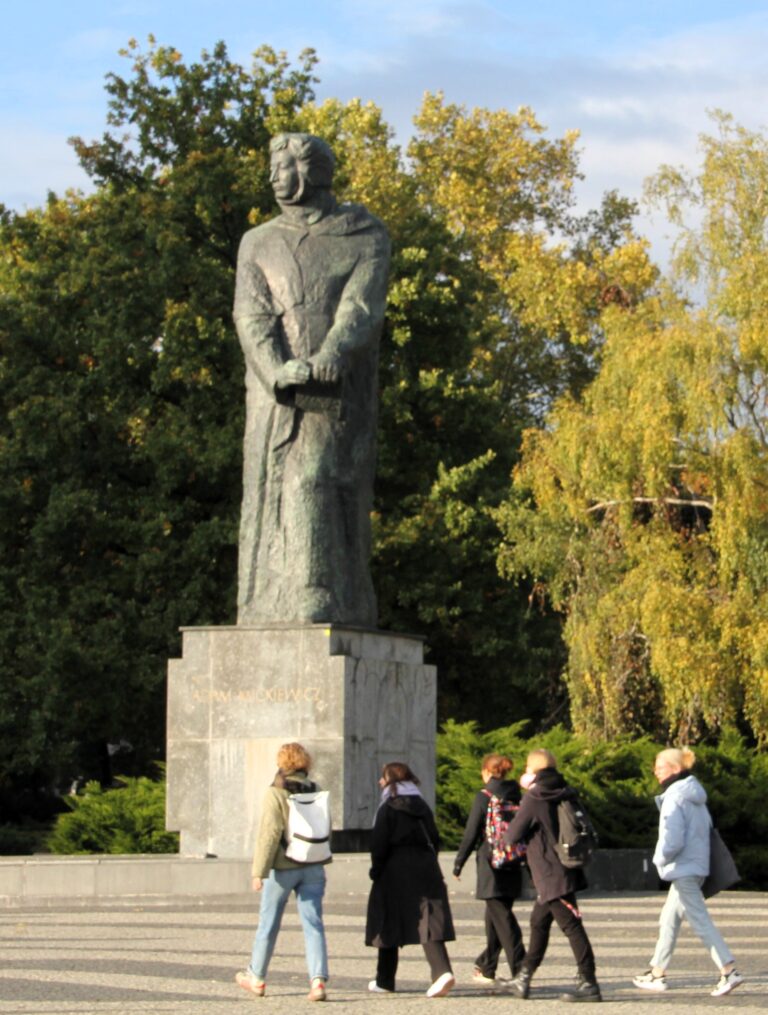 Wysoki pomnik Adama Mickiewicza w Poznaniu. Wykonany jest z kamienia i przedstawia całą postać poety. Pod pomnikiem przechodzą ludzie. W tle drzewa