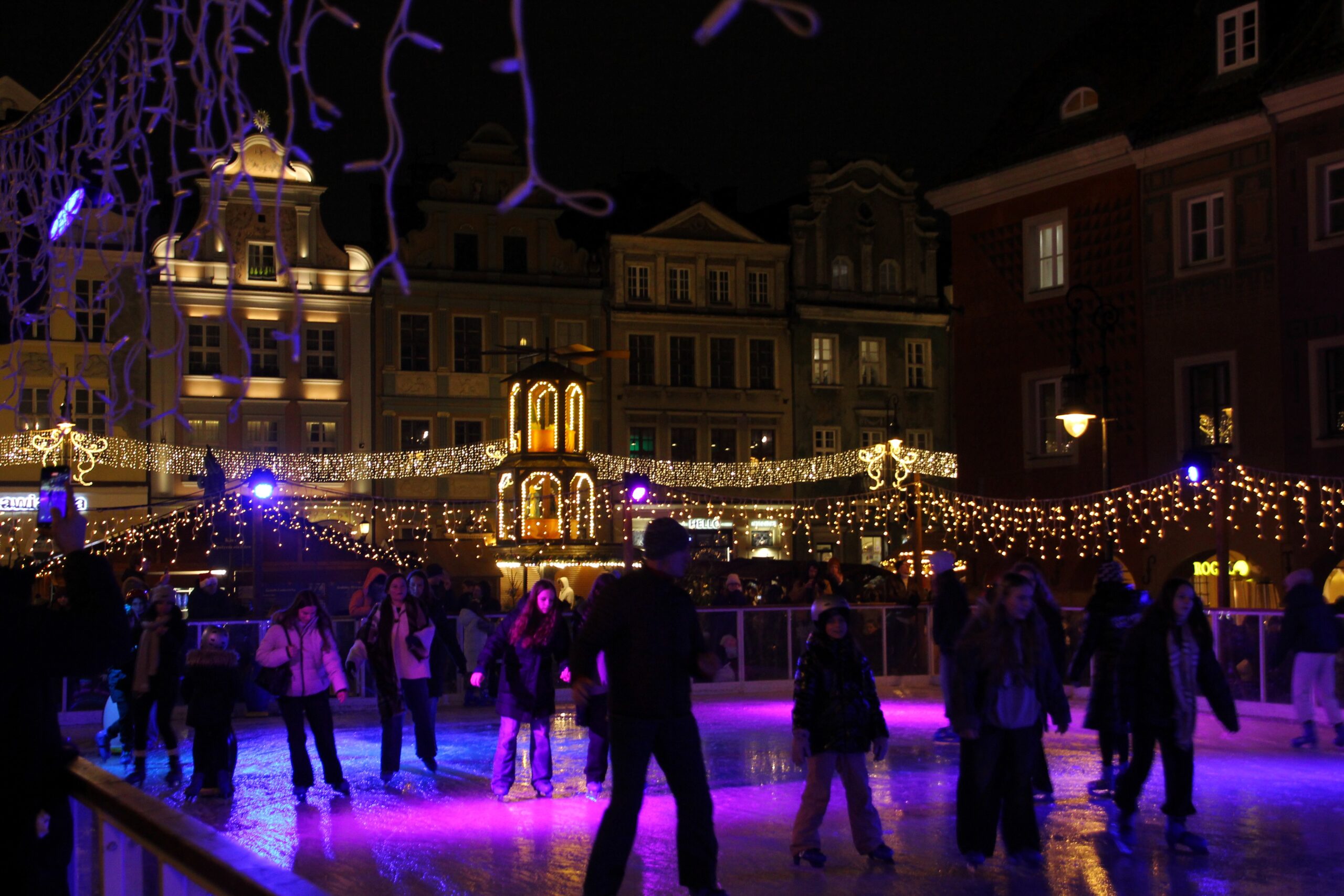 Ludzie na lodowisku, w tle świąteczne oświetlenia jarmarku i kamienice