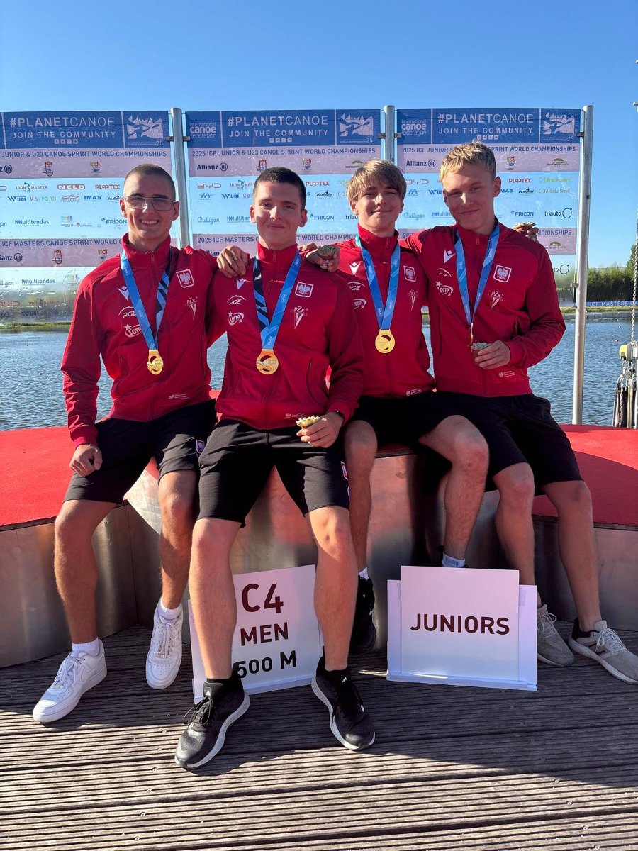 Kacper Stanek ze złotym medalem MŚ juniorów w kajakarstwie. Zawodnik jest w otoczeniu 3 kolegów, siedzą na podium w czerwonych bluzach reprezentacji Polski, z medalami na szyi. W tle zbiornik wodny.