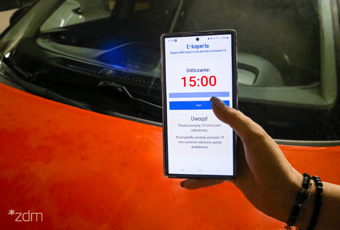 Osoba trzyma telefon z uruchomioną aplikacją do odmierzania czasu postoju na kopercie, a w tle widoczna jest pomarańczowa maska samochodu.