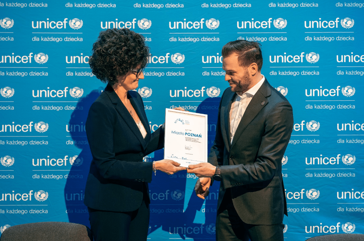 Dwie osoby w eleganckich strojach stoją przed ścianką UNICEF i trzymają wspólnie dokument.