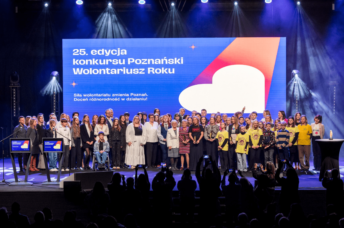 Duża grupa osób stoi na scenie, a za nimi widoczny jest duży ekran z napisem konkursu na niebieskim tle. Na ekranie znajduje się białe serce z ciągnącą się fioletowo-pomarańczową smugą.