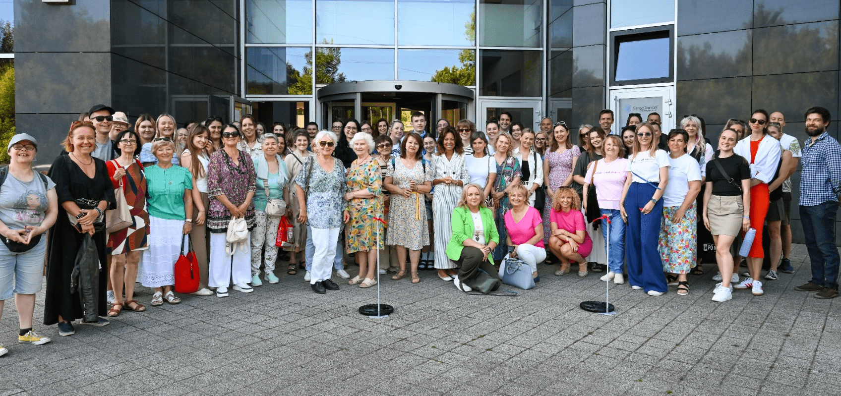 Duża grupa ludzi stoi razem i pozuje do zdjęcia przed oszklonym wejściem do budynku.