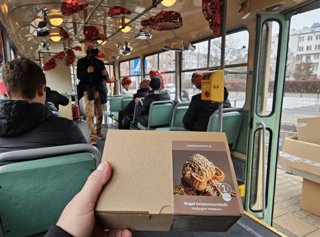 Osoba trzyma karton z rogalem w środku tramwaju, w którym siedzą pasażerowie zwróceni w stronę stojącej osoby. Przy suficie tramwaju widoczne są czerwono-czarne dekoracje.