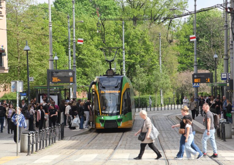 Tramwaj na przystanku, dookoła dużo ludzi czekających, kilku pieszych przechodzi przez pasy, w tle zieleń
