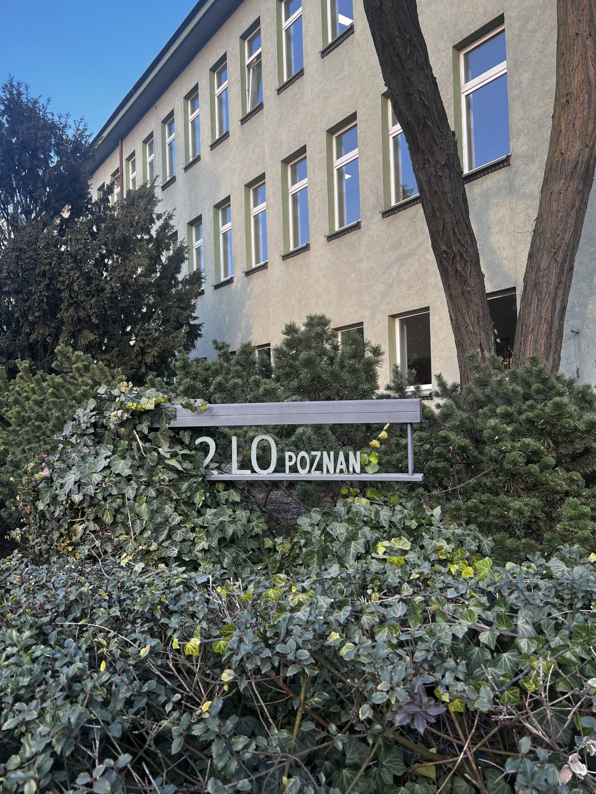 Fragment szarego, dwupiętrowego budynku szkoły z rzędami okien. Na pierwszym planie, wśród gęstych krzewów i pnączy, znajduje się metalowa tablica z napisem „2 LO POZNAŃ”. Po prawej stronie rosną wysokie drzewa.
