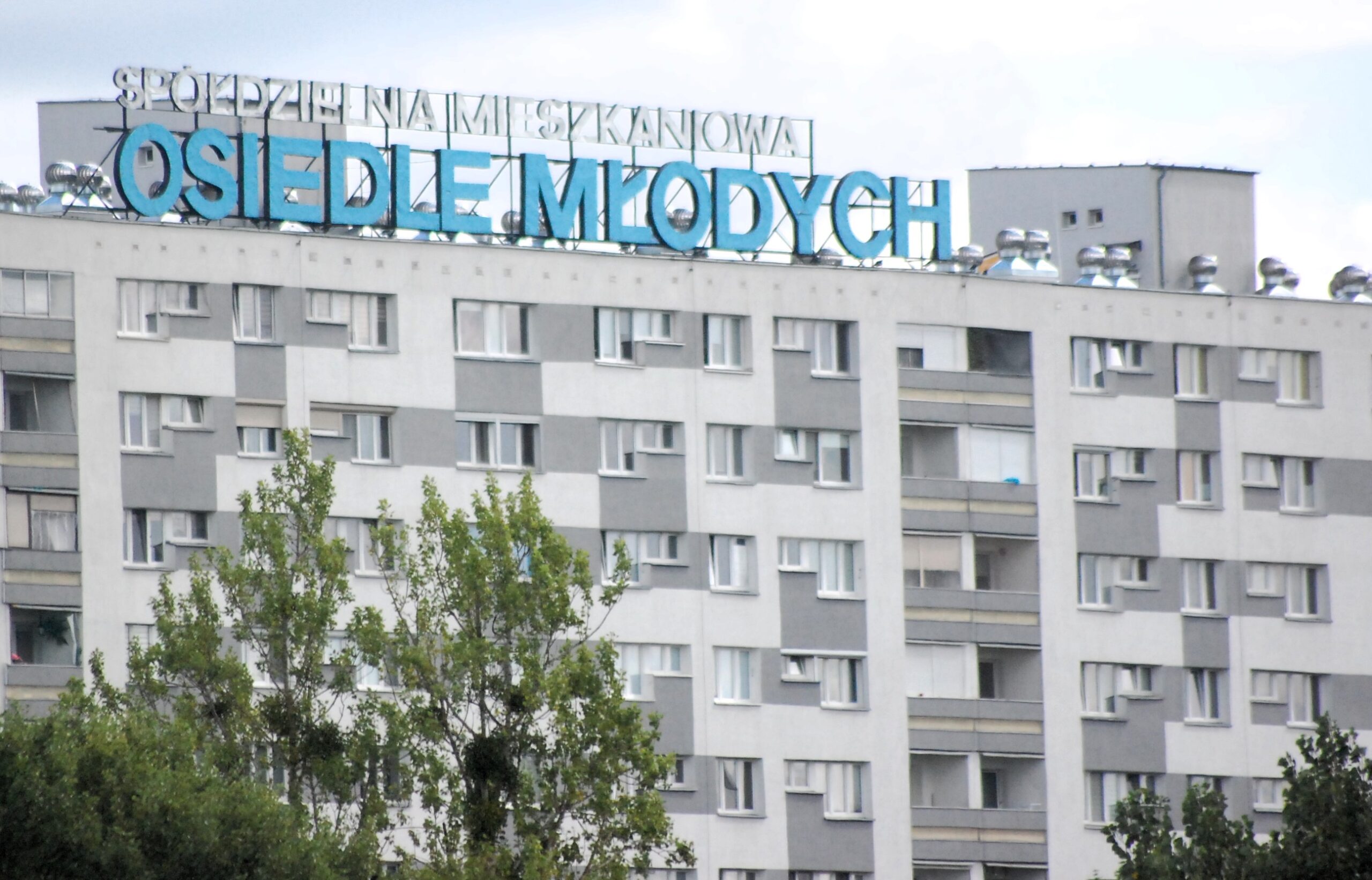 Wysoki blok z wielkiej płyty z dużym napisem "Osiedle Młodych". Blok jest płaski, ma loggie, przed nim zieleń.